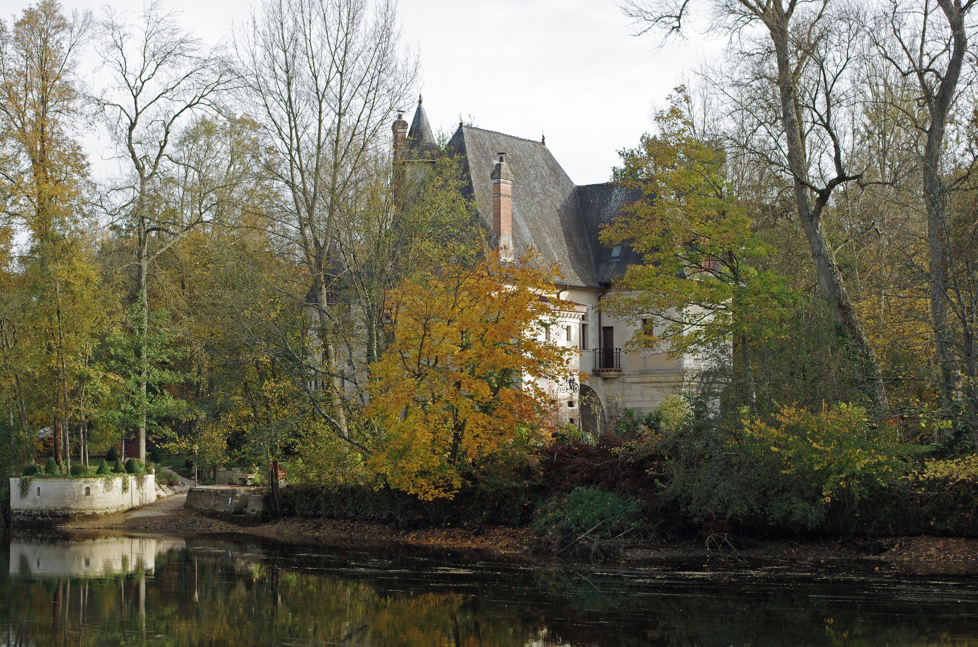 A historic building with a steep roof and a prominent chimney, surrounded by autumn-colored trees and a calm river.