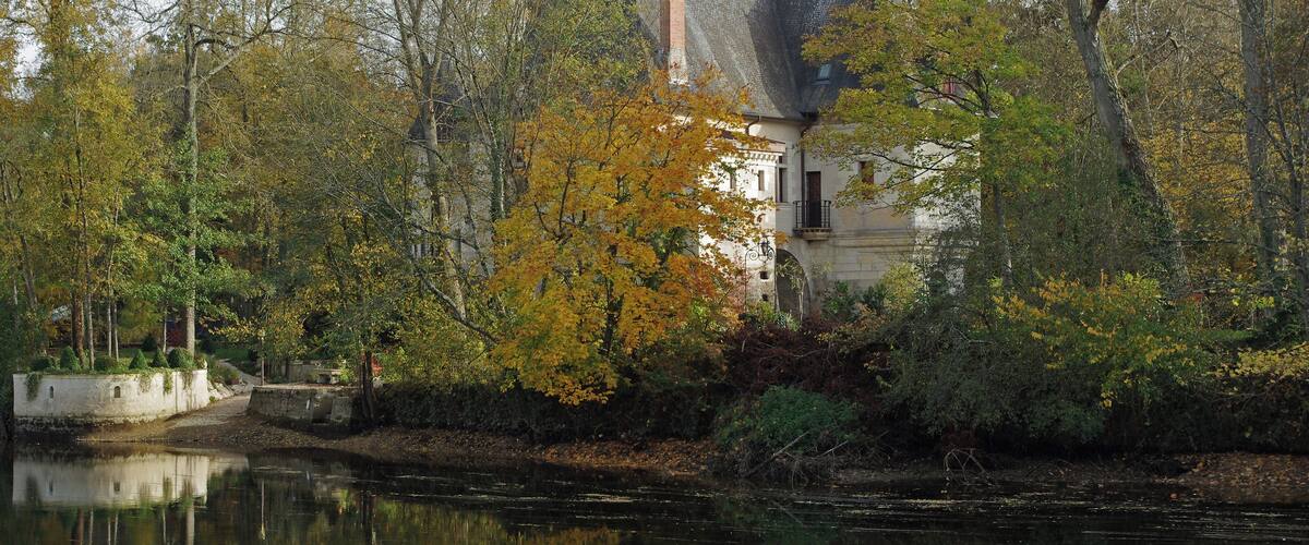 A historic building with a steep roof and a prominent chimney, surrounded by autumn-colored trees and a calm river.