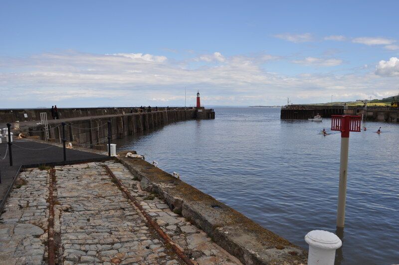 Un muelle de piedra que se adentra en aguas tranquilas, un faro a lo lejos y un cielo despejado.