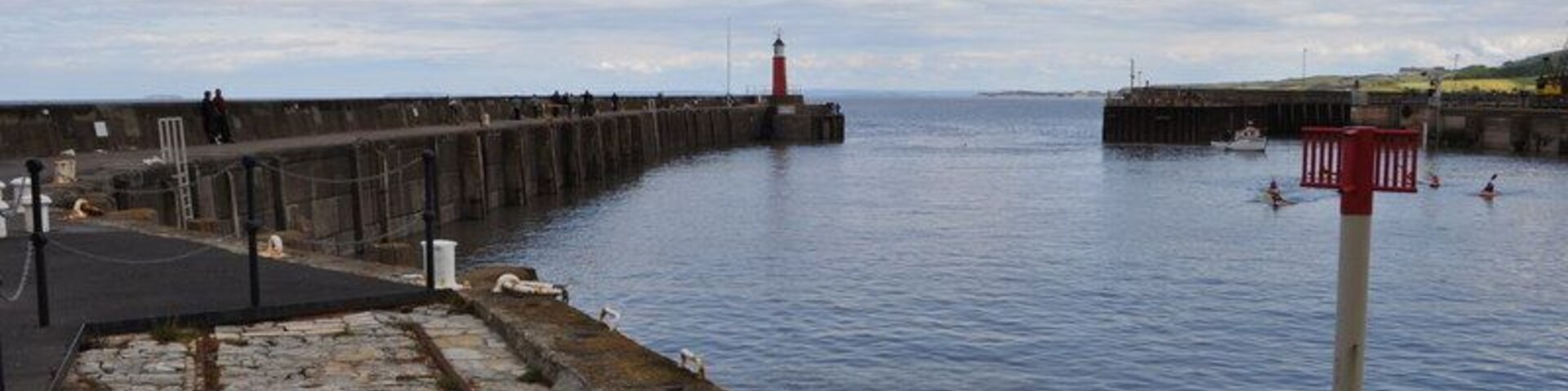 Un muelle de piedra que se adentra en aguas tranquilas, un faro a lo lejos y un cielo despejado.