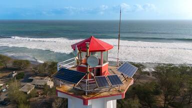 The lighthouse stands tall with solar panels, overlooking the crashing ocean waves on a clear day