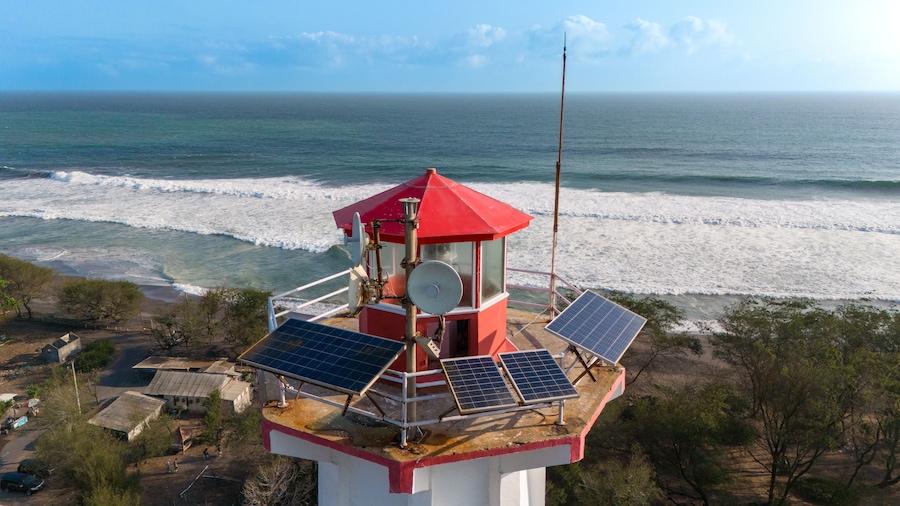 The lighthouse stands tall with solar panels, overlooking the crashing ocean waves on a clear day