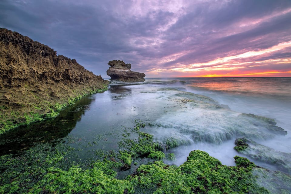 A coastal landscape with a prominent rock formation, green algae-covered rocks, and a sunset over the ocean.