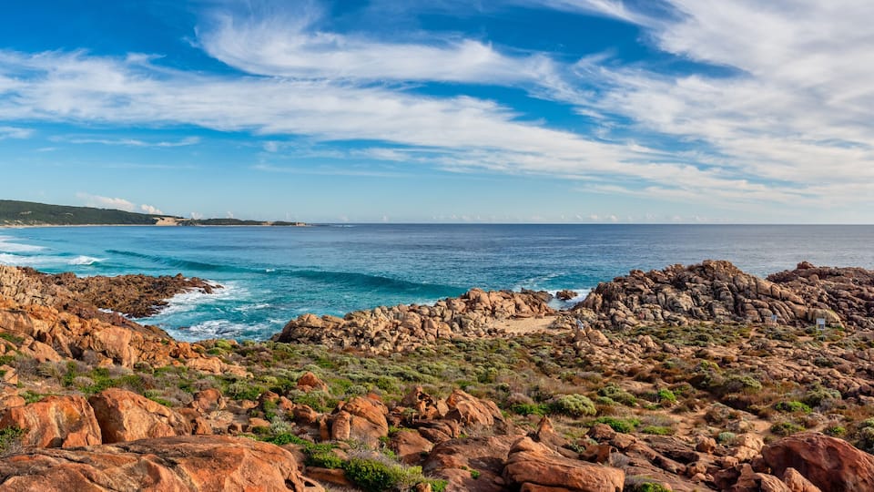 A rocky coastline with waves crashing against the shore, a clear blue sky, and distant wind turbines.