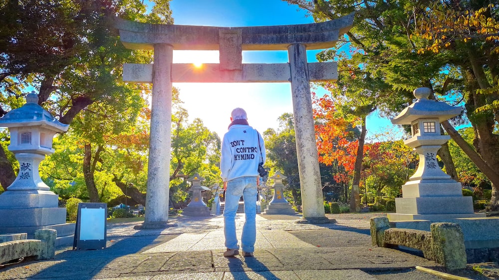 A person walking towards a torii gate in a park with stone lanterns and trees.
