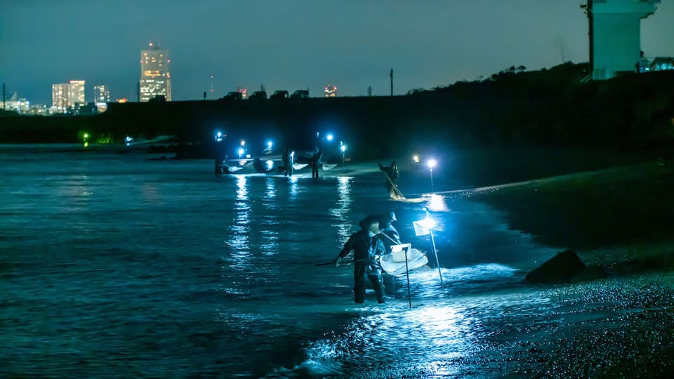 A nighttime scene with a river reflecting city lights, a person wading in the water, and a distant city skyline.