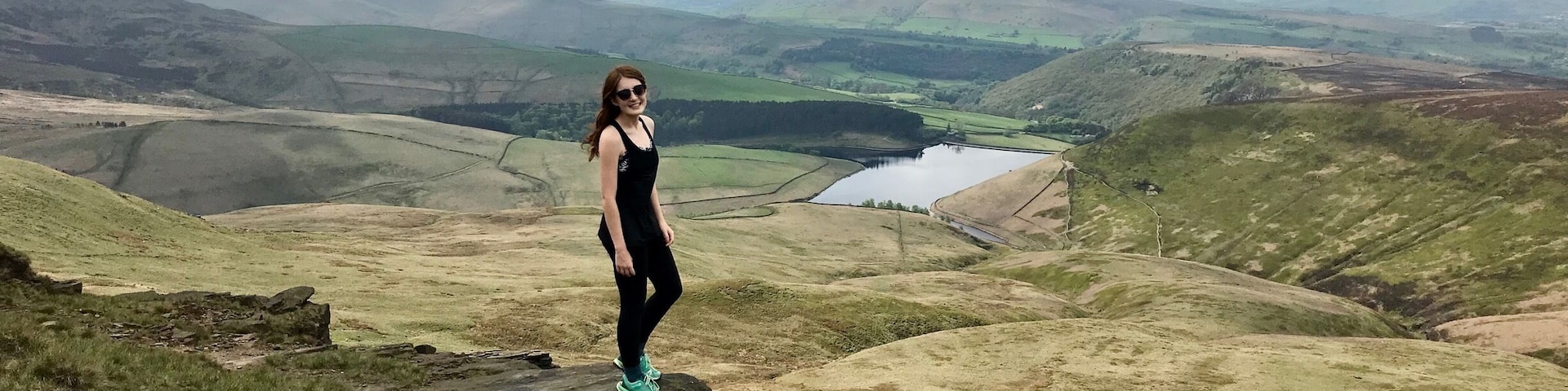 A person standing on a rock with a view of rolling hills and a lake in the distance.