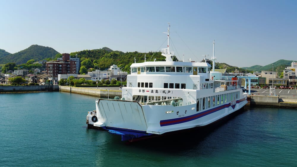 A large white ferry docked at a pier with a cityscape and mountains in the background.