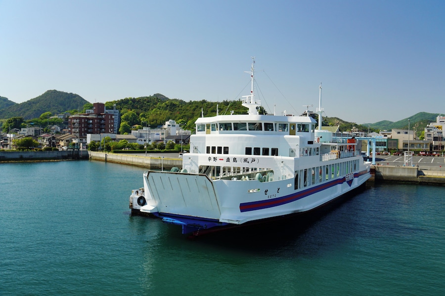 Un gran ferry blanco atracado en un muelle, con un paisaje urbano y montañas al fondo.