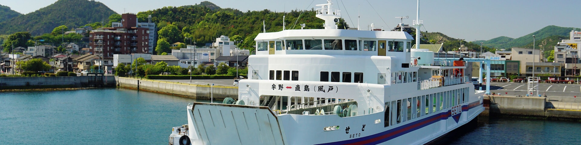 Un gran ferry blanco atracado en un muelle, con un paisaje urbano y montañas al fondo.