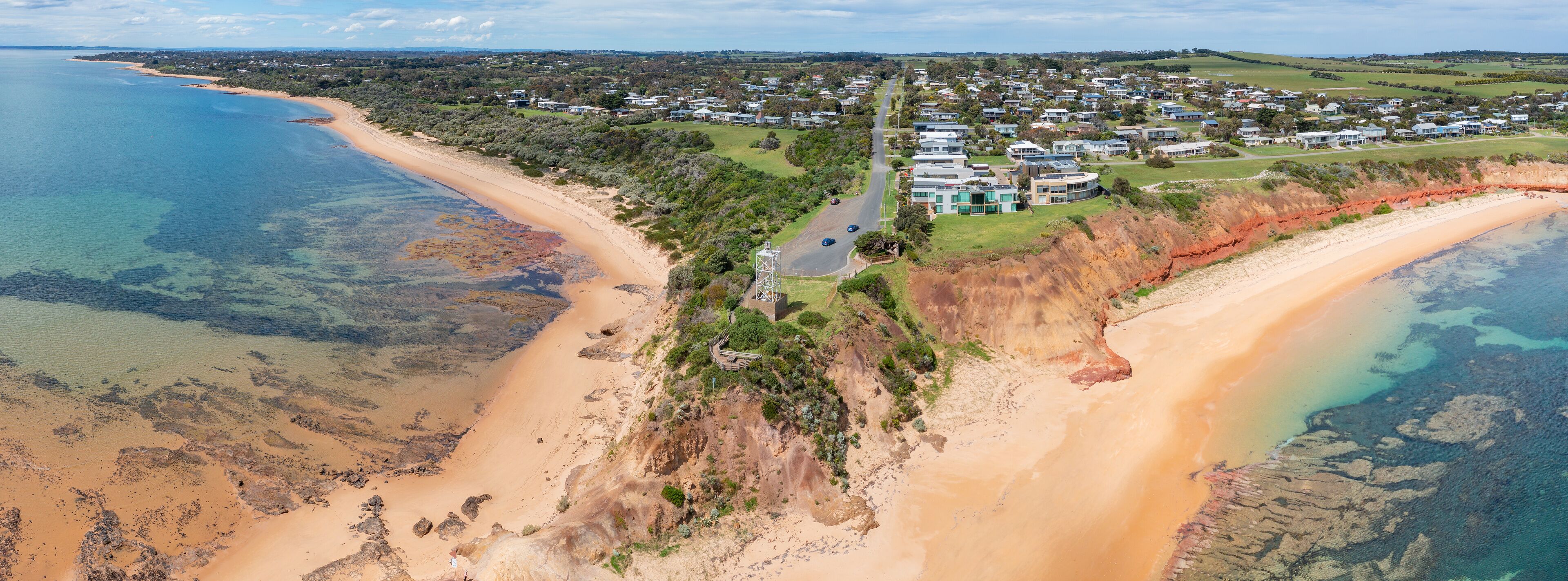 An aerial view of a coastal town with a beach, houses, and a road leading to a hilltop with a viewing platform.