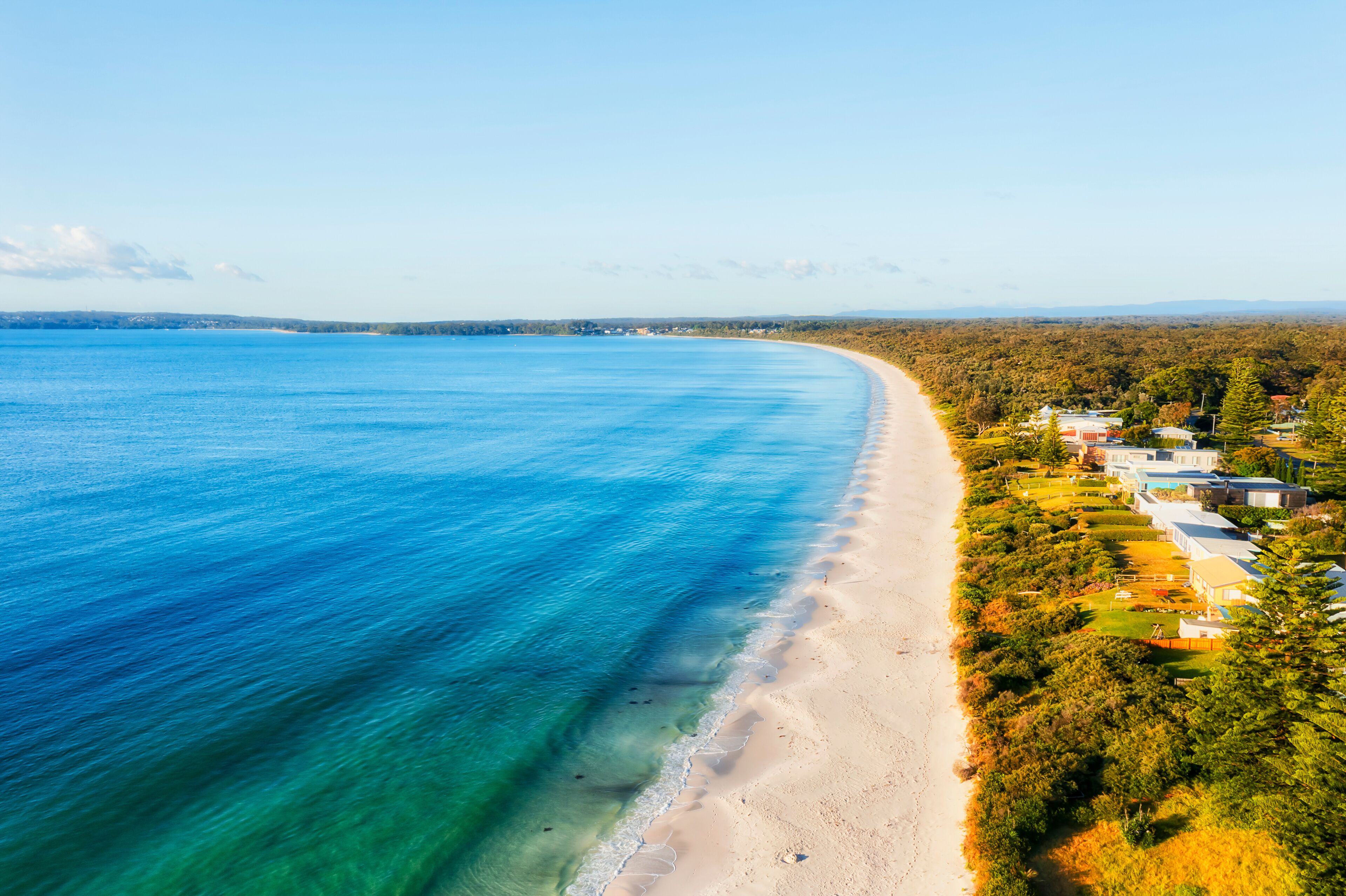 A coastal landscape with a sandy beach, clear blue waters, and a residential area with houses and greenery.