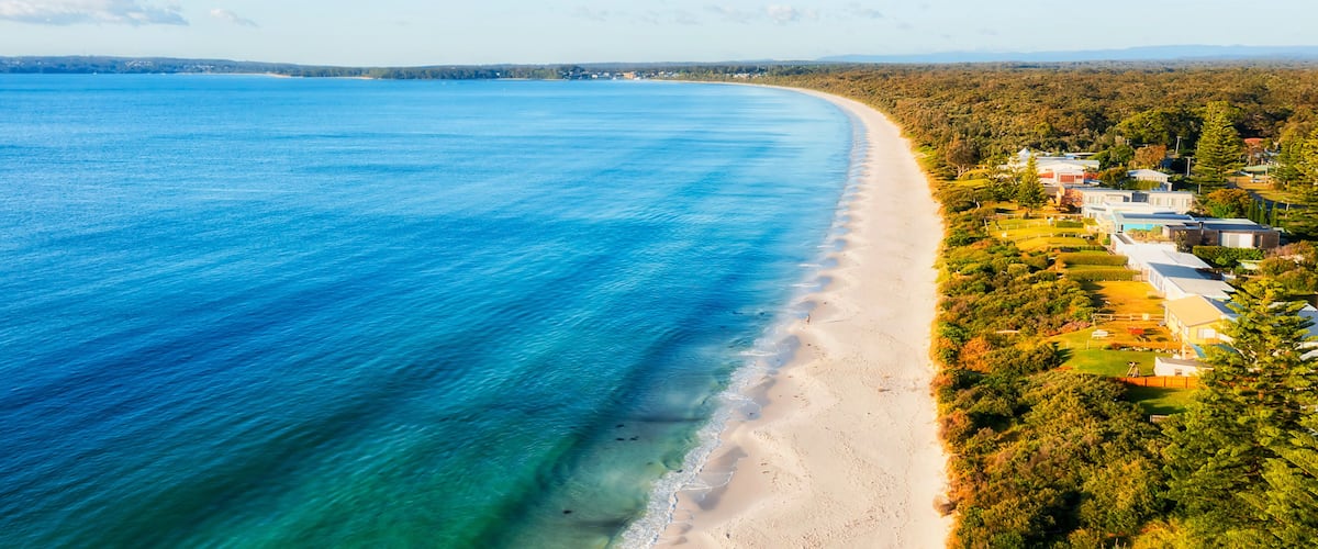 A coastal landscape with a sandy beach, clear blue waters, and a residential area with houses and greenery.