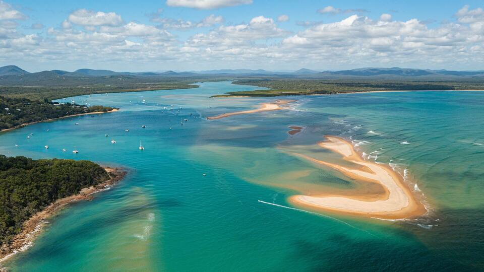 An aerial view of a coastal area with clear turquoise waters, a sandy beach, and a backdrop of distant mountains.