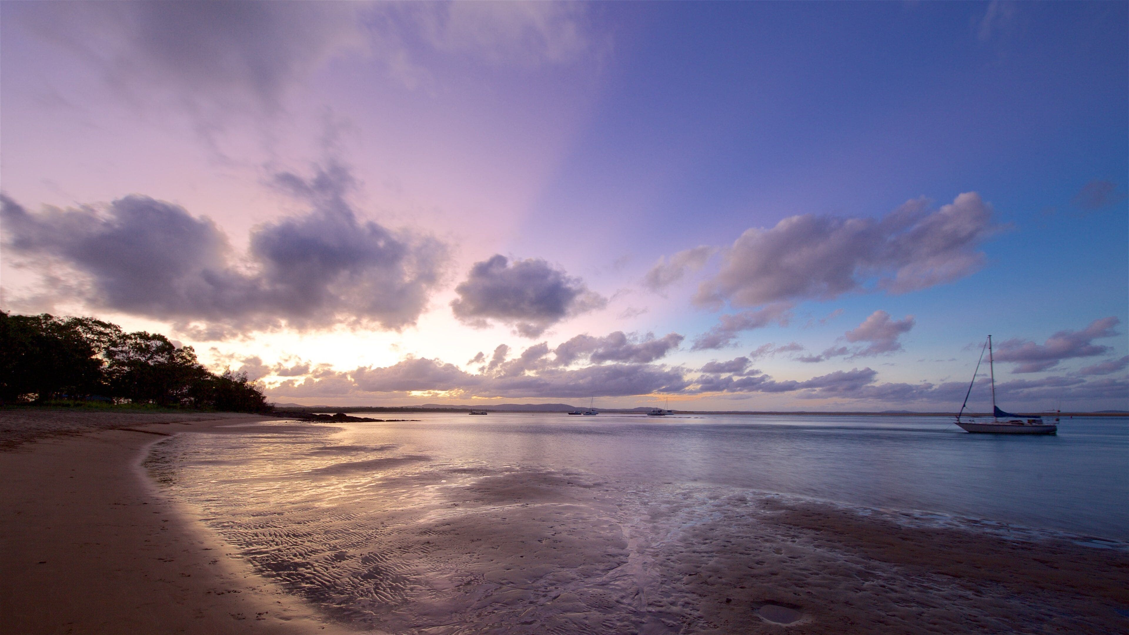 A beach with a sailboat, calm water, and a sunset sky.