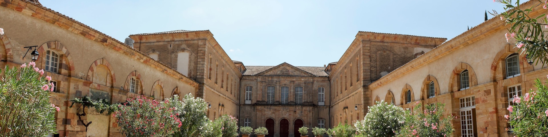 Ein Innenhof mit einem zentralen Gebäude, das von zwei Flügeln gesäumt wird, umgeben von Blumentöpfen und blauem Himmel.