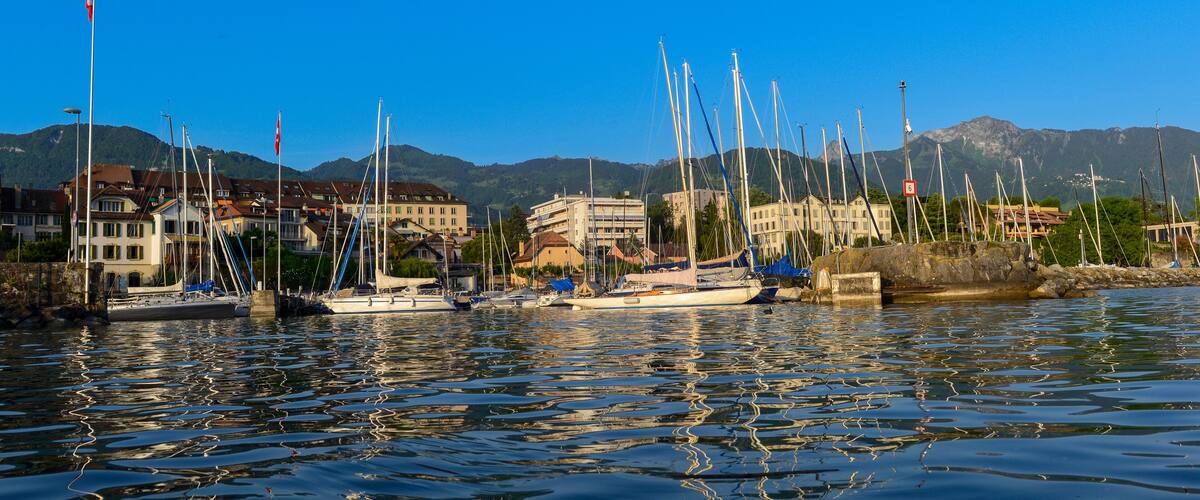 A marina with sailboats docked, buildings, and mountains in the background.