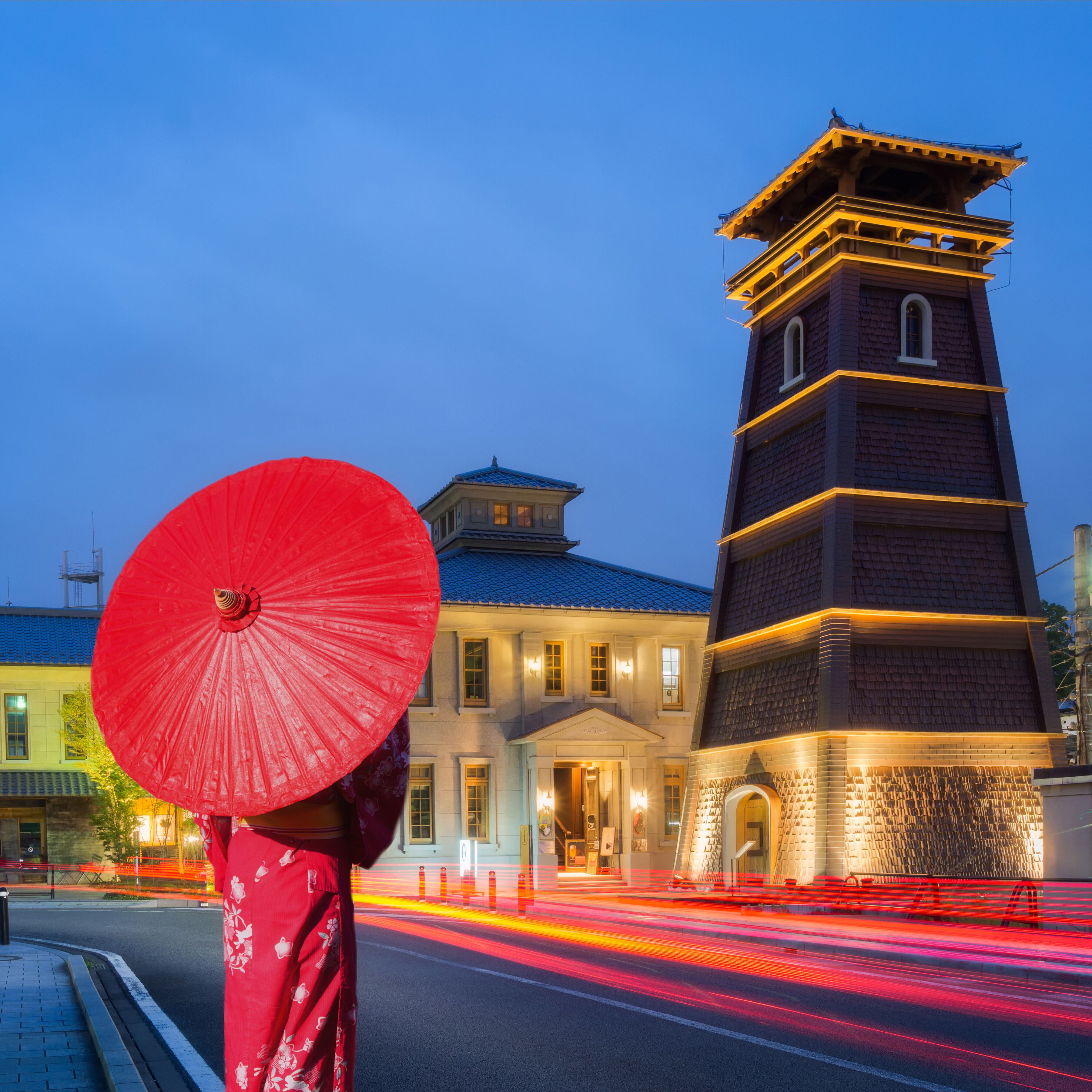 A person holding a red umbrella, a traditional building with a tower, and a street with light trails from moving vehicles.