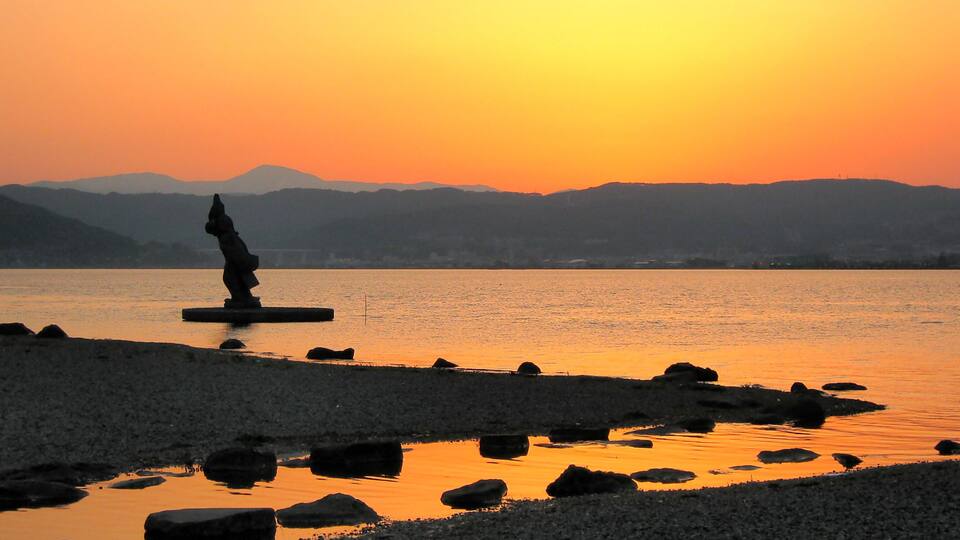 A statue on a rock by a calm lake at sunset with mountains in the background.