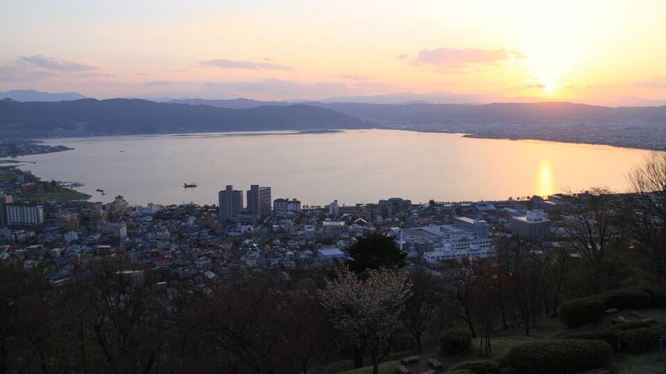 A cityscape with a large body of water, mountains in the background, and a sunset.