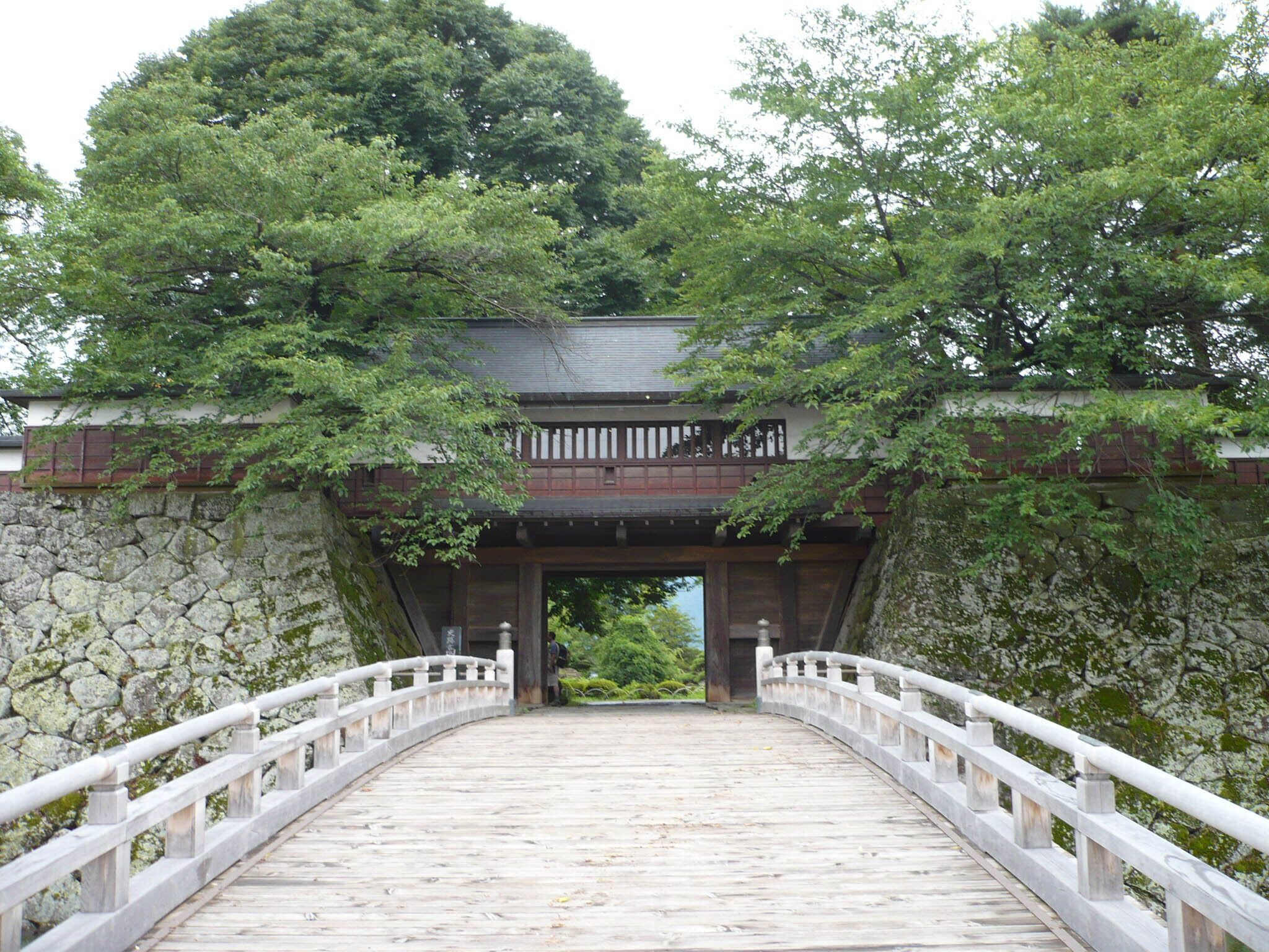 A stone bridge with a wooden walkway leading to a traditional Japanese building under a tree canopy.