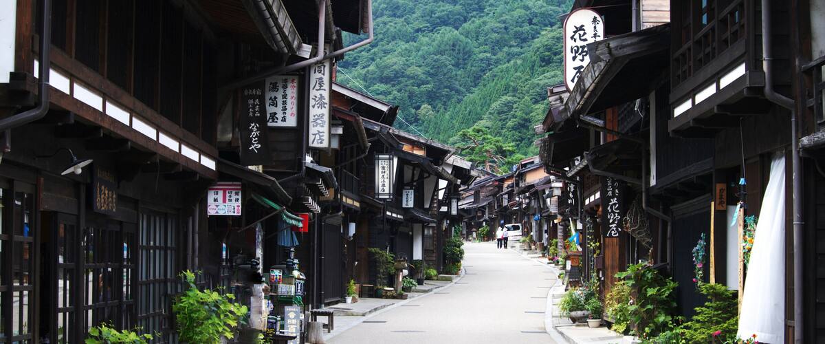 A narrow street lined with traditional wooden buildings, potted plants, and signs in a foreign language.