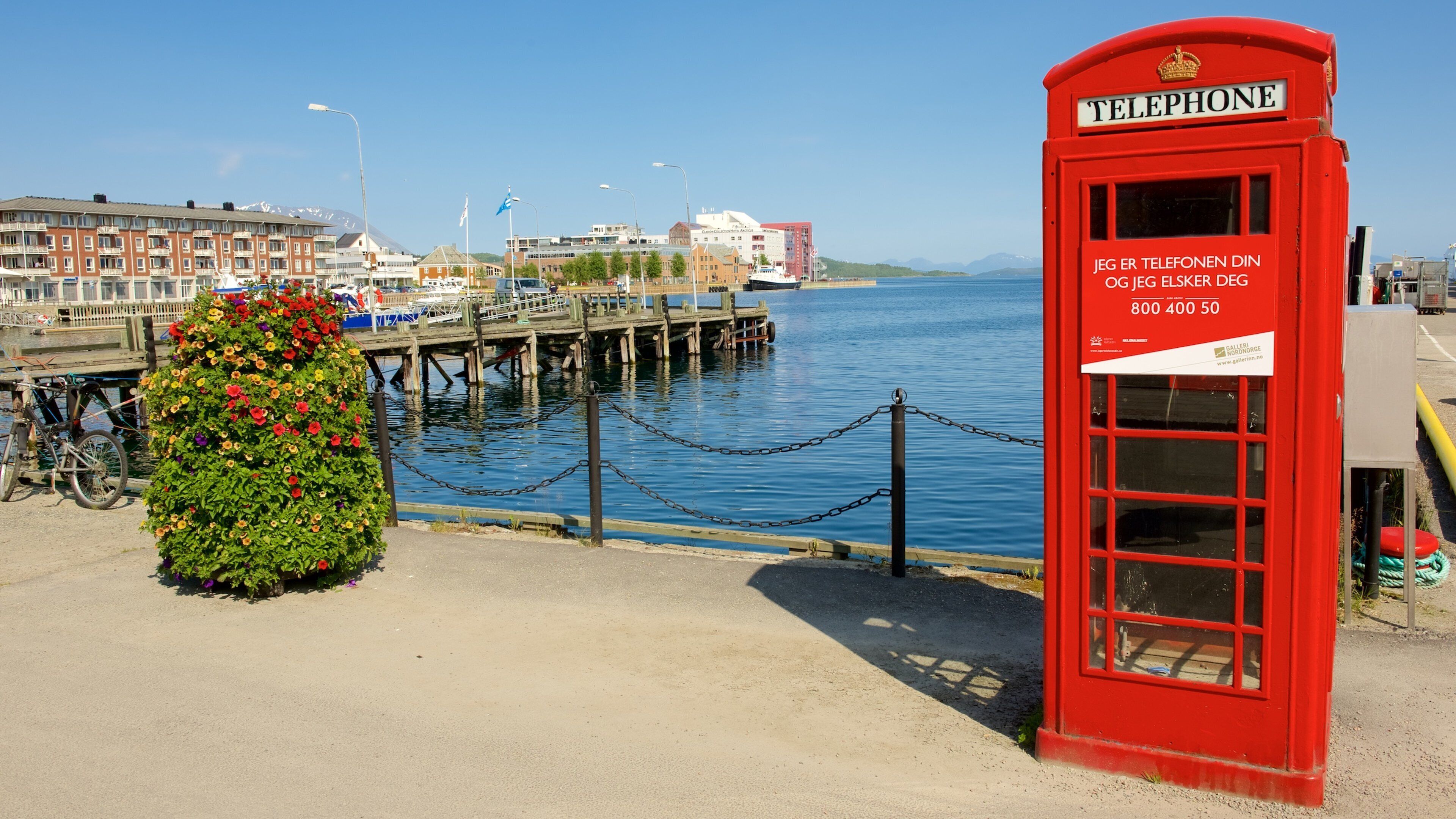 A red telephone booth with a sign in a foreign language, a flower bed, a dock, and a body of water.