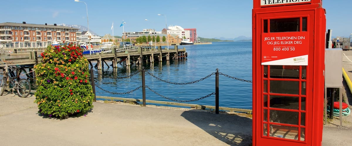 A red telephone booth with a sign in a foreign language, a flower bed, a dock, and a body of water.