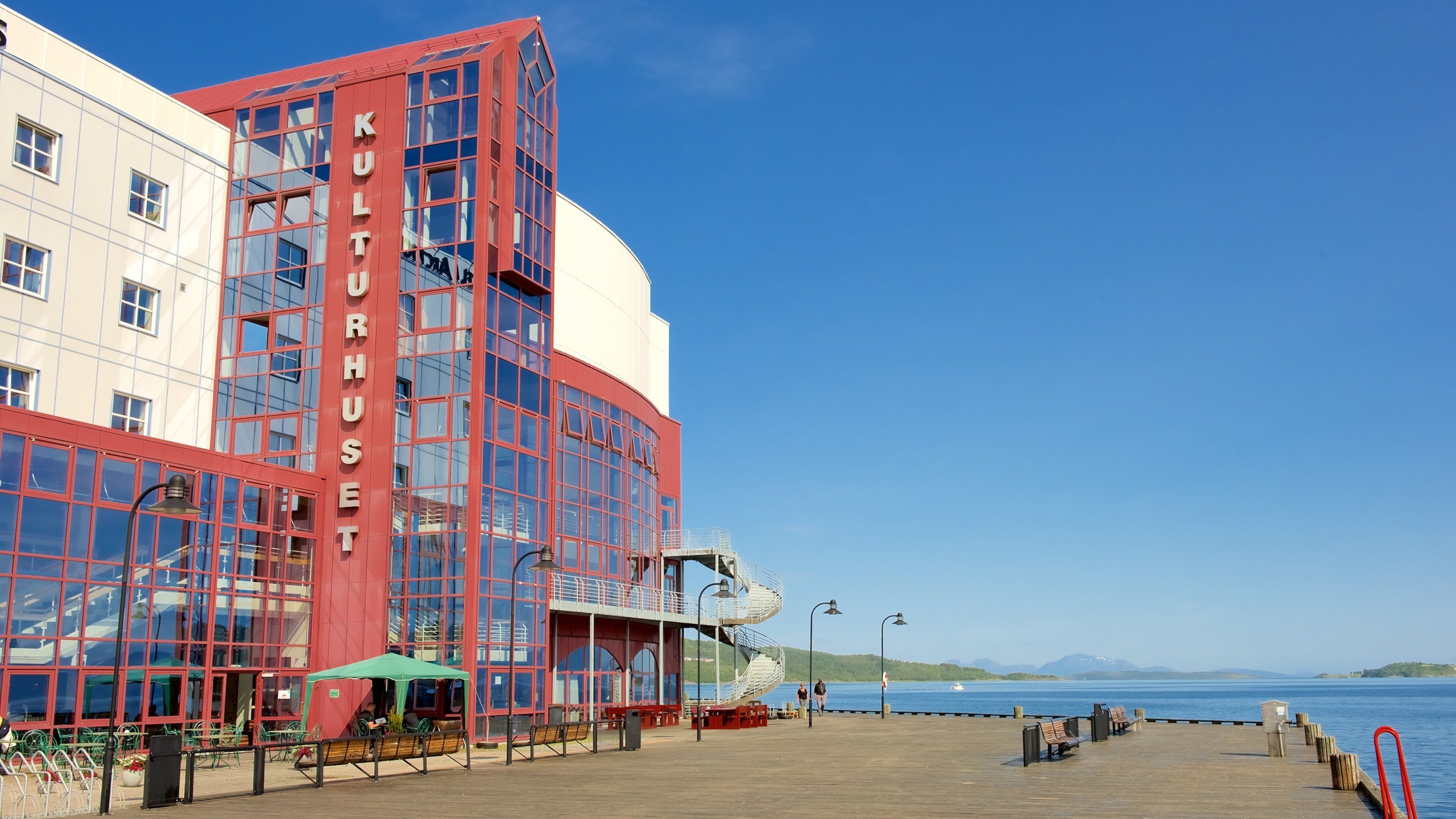 Un edificio sul lungomare con facciata rossa e ampie finestre, un molo di legno con panchine, e un cielo azzurro limpido.