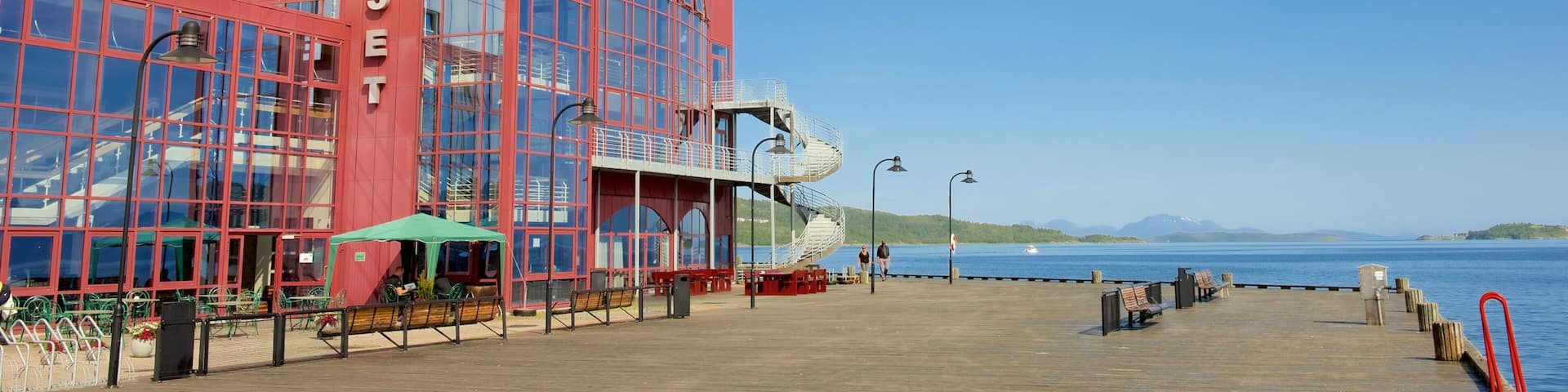 A waterfront building with a red facade and large windows, a wooden pier with benches, and a clear blue sky.