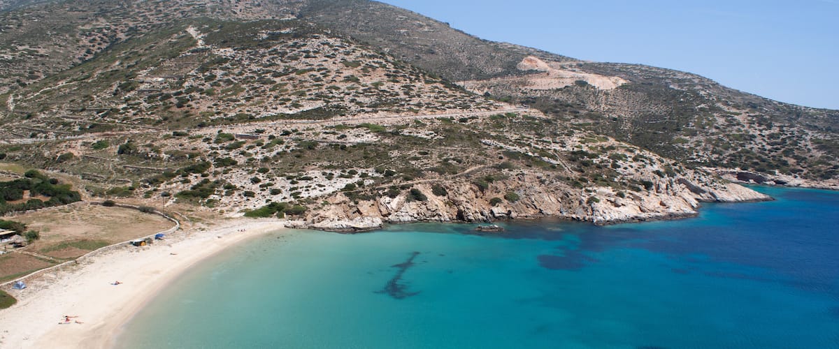 A coastal landscape with a sandy beach, clear turquoise waters, rocky cliffs, and sparse vegetation.