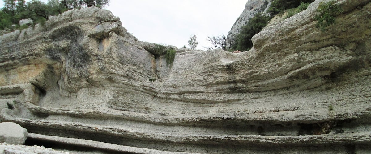 A rocky cliff with layered sedimentary rock formations and a small pond at its base.