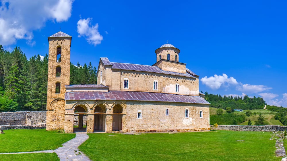 Une église historique en pierre, dotée d’un clocher et d’une coupole, entourée de champs verdoyants et d’arbres luxuriants.