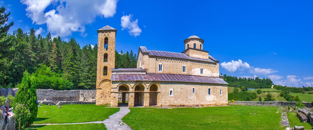 A historic stone church with a bell tower and a dome, surrounded by lush green fields and trees.