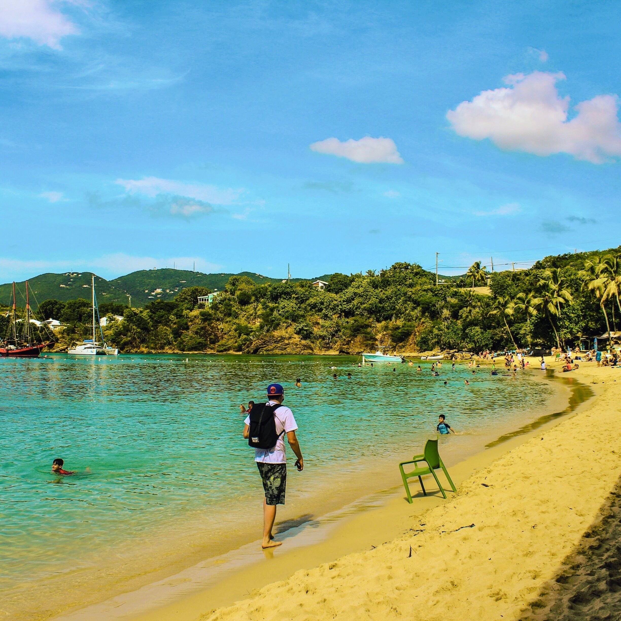 A beach with clear turquoise water, a person walking towards the water, boats, and a green chair on the sand.