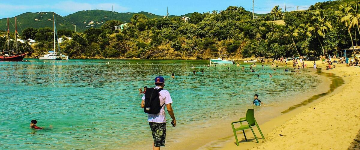 A beach with clear turquoise water, a person walking towards the water, boats, and a green chair on the sand.