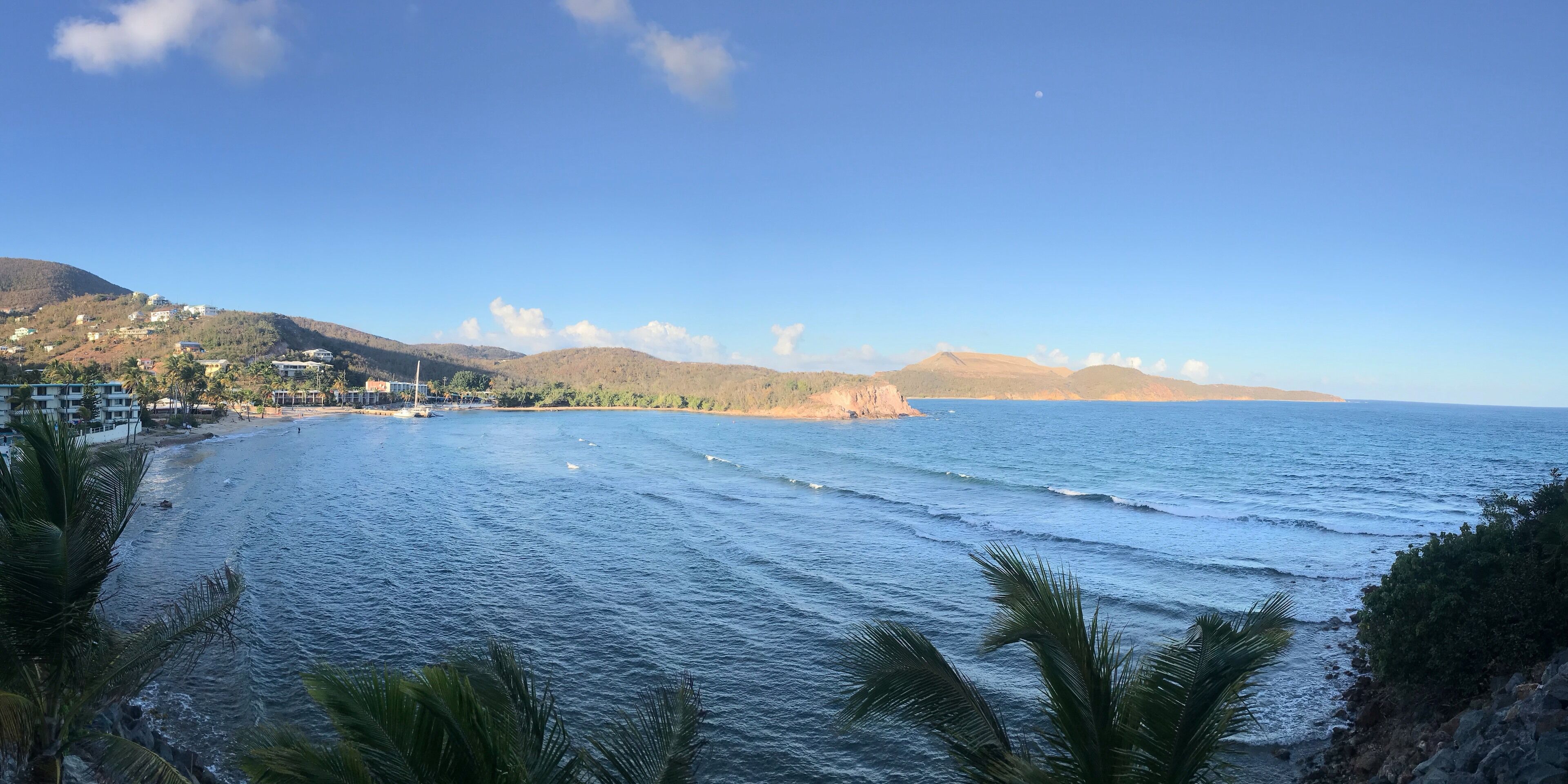 A coastal landscape with a beach, ocean waves, palm trees, and distant hills.