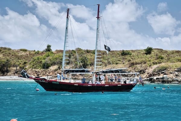 Ein Segelboot mit zwei Masten auf einem türkisblauen Meer, umgeben von felsiger Küste und vereinzelter Vegetation.