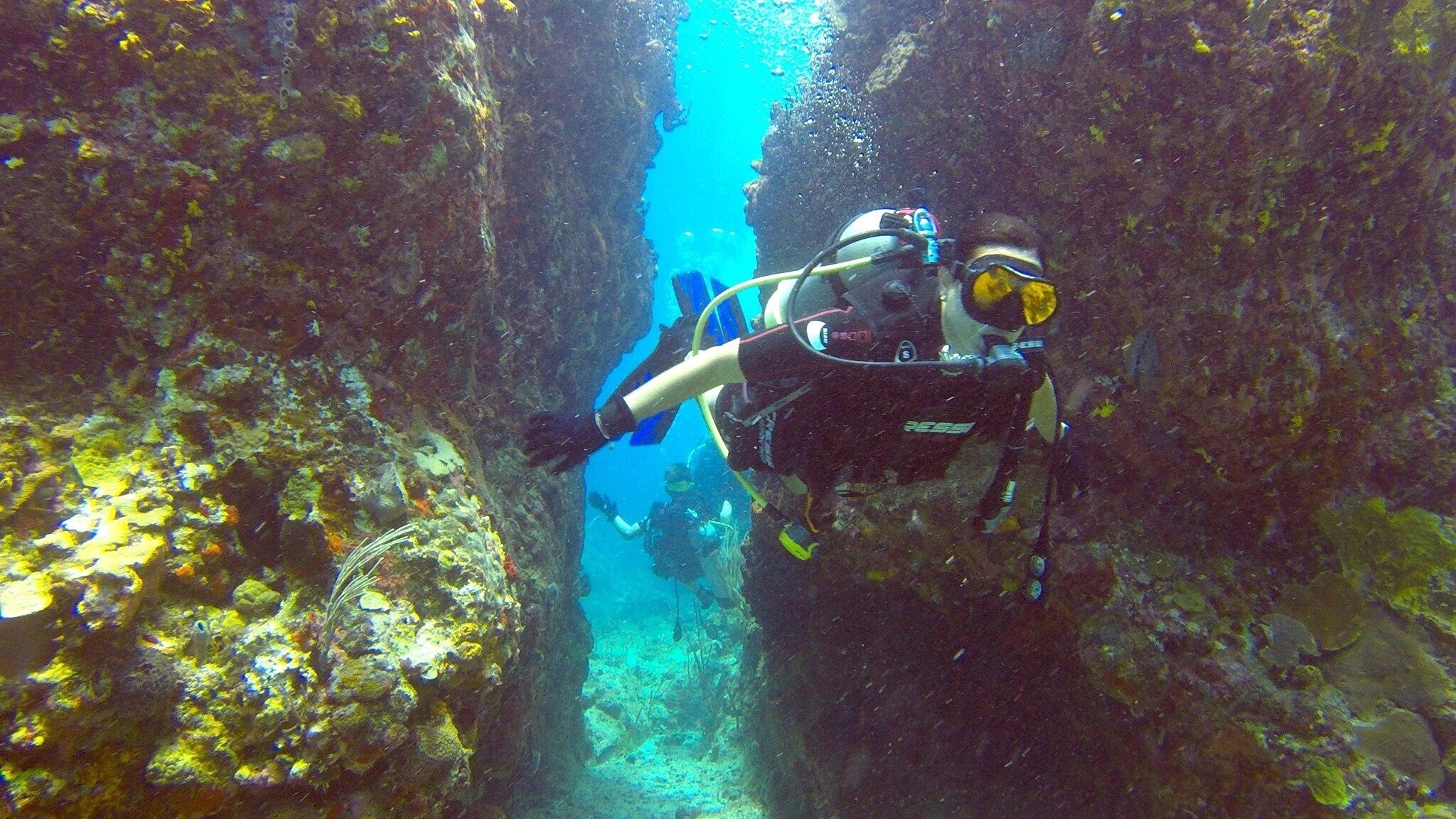 Mergulhador em uma passagem subaquática estreita, cercado por formações rochosas e vida marinha.