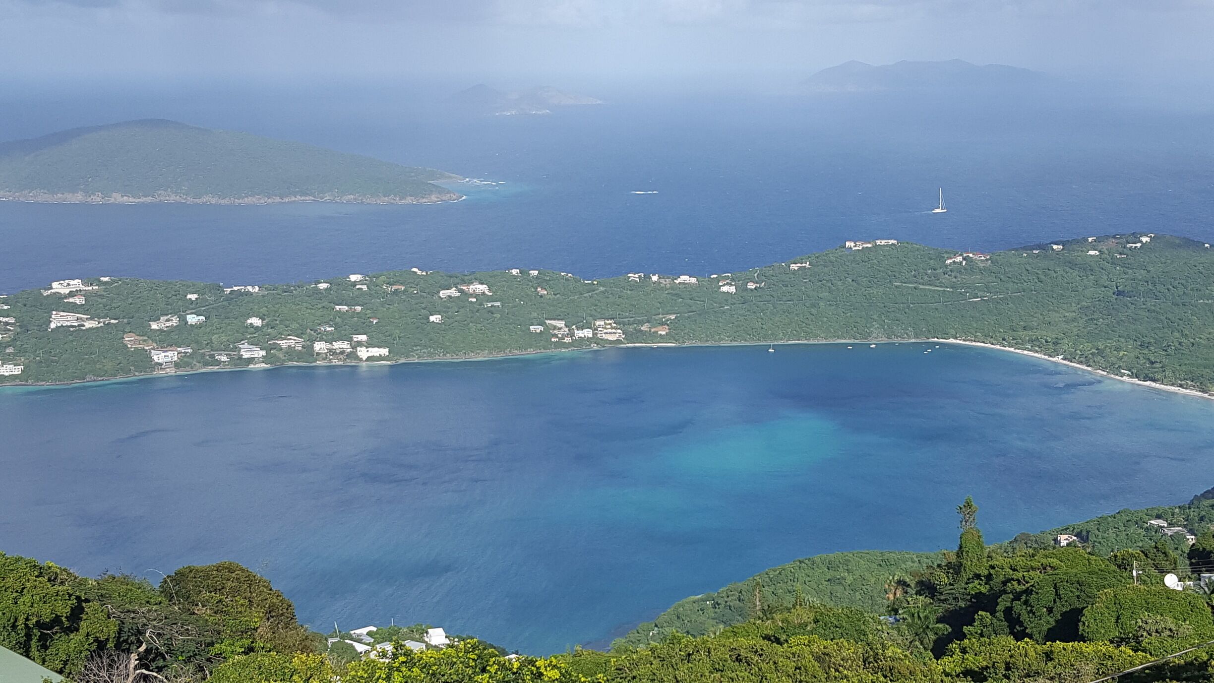 A coastal landscape with a clear blue sea, green hills, and scattered buildings.