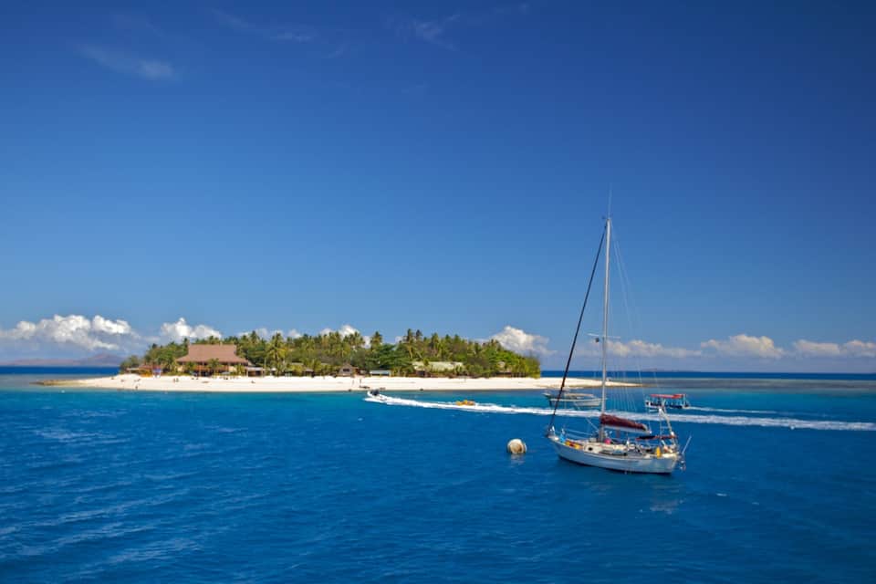 Mar azul cristalina con un velero, una playa arenosa y una pequeña isla con edificios y árboles.