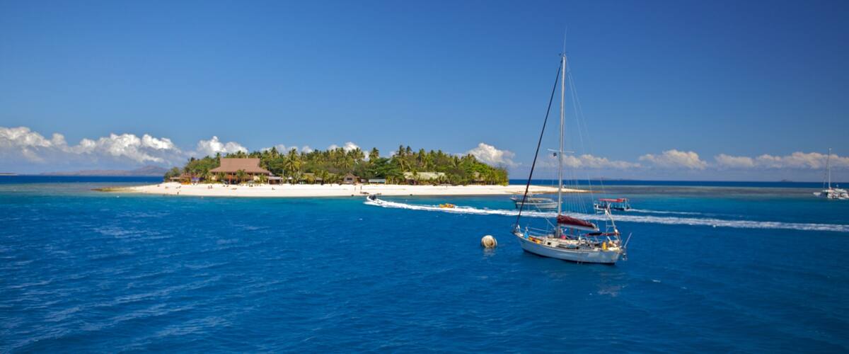 A clear blue sea with a sailboat, a sandy beach, and a small island with buildings and trees.