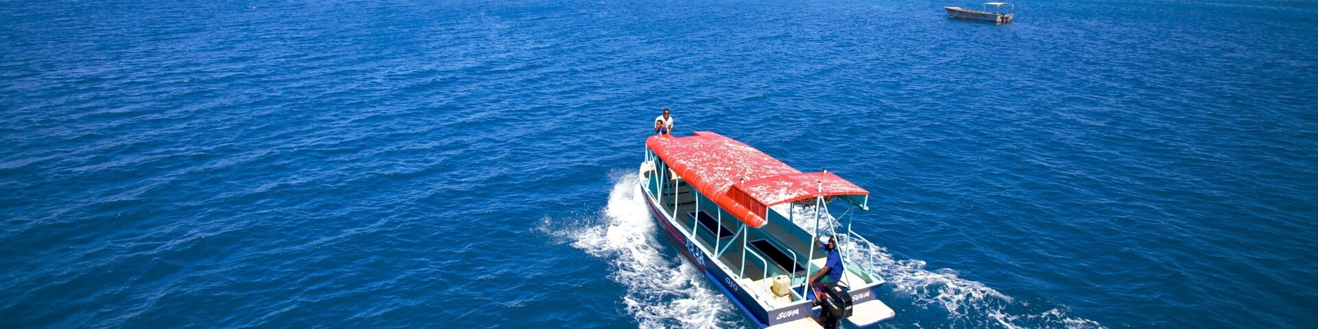 Un barco con toldo rojo navegando por el océano, una pequeña isla con palmeras al fondo y otro barco a lo lejos.