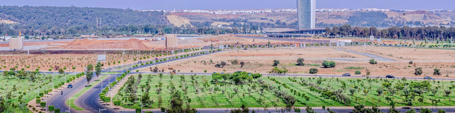 A modern skyscraper, a highway, and a hilly landscape with sparse vegetation.