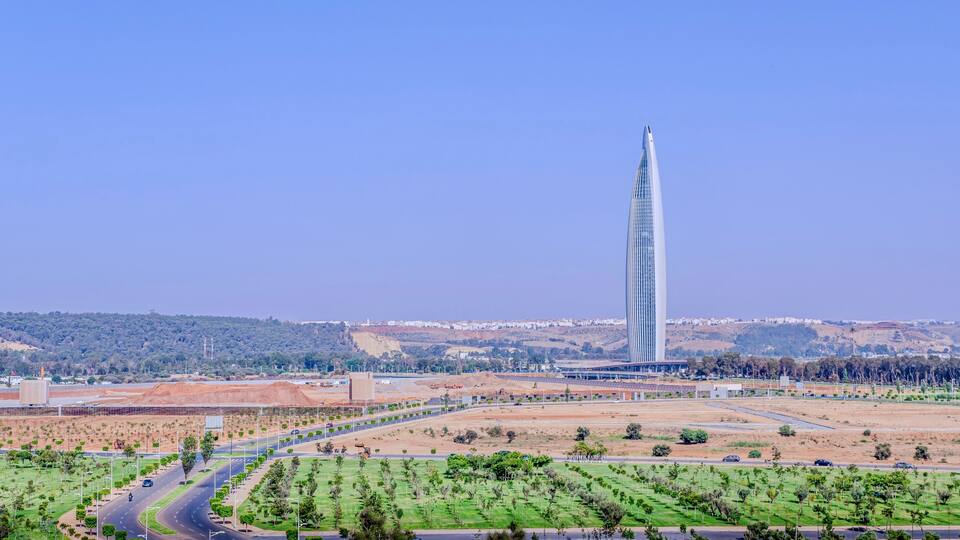 A modern skyscraper, a highway, and a hilly landscape with sparse vegetation.