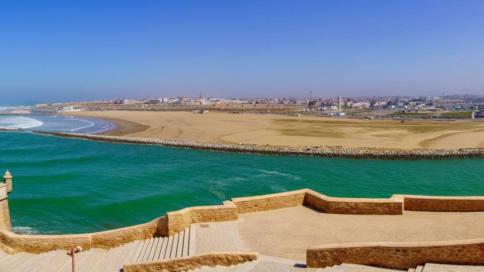 A coastal city with a sandy beach, a stone sea wall, and a clear blue sky.