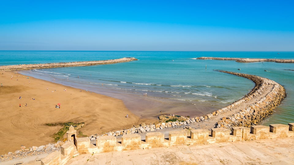 A coastal beach with a stone seawall, clear blue water, and sandy shore.