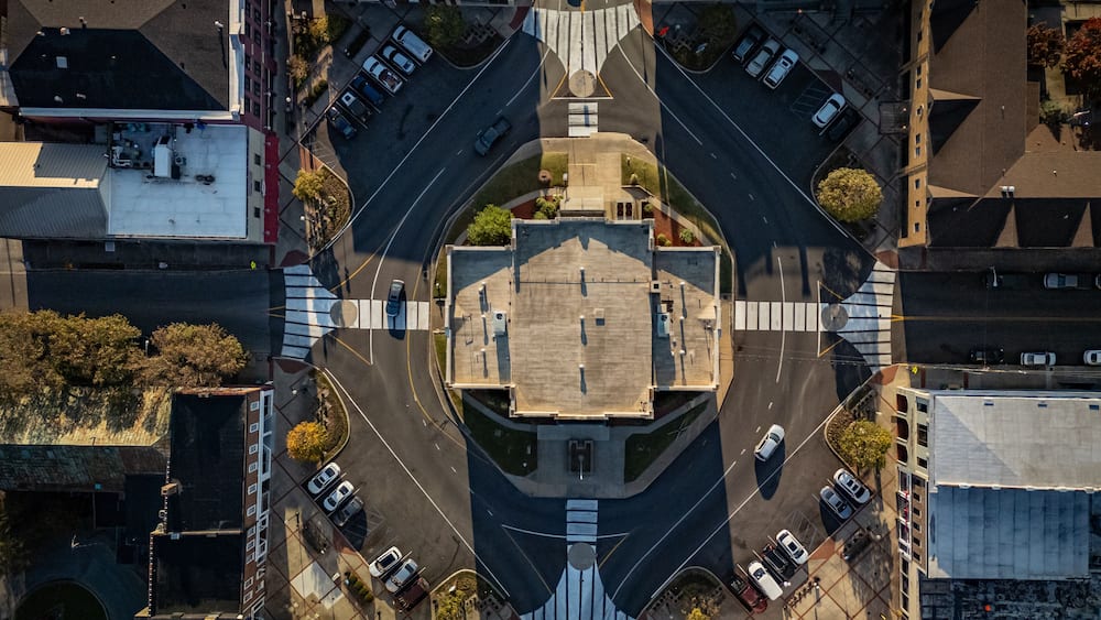 An aerial view of a roundabout with a central plaza, surrounded by buildings and parked cars.