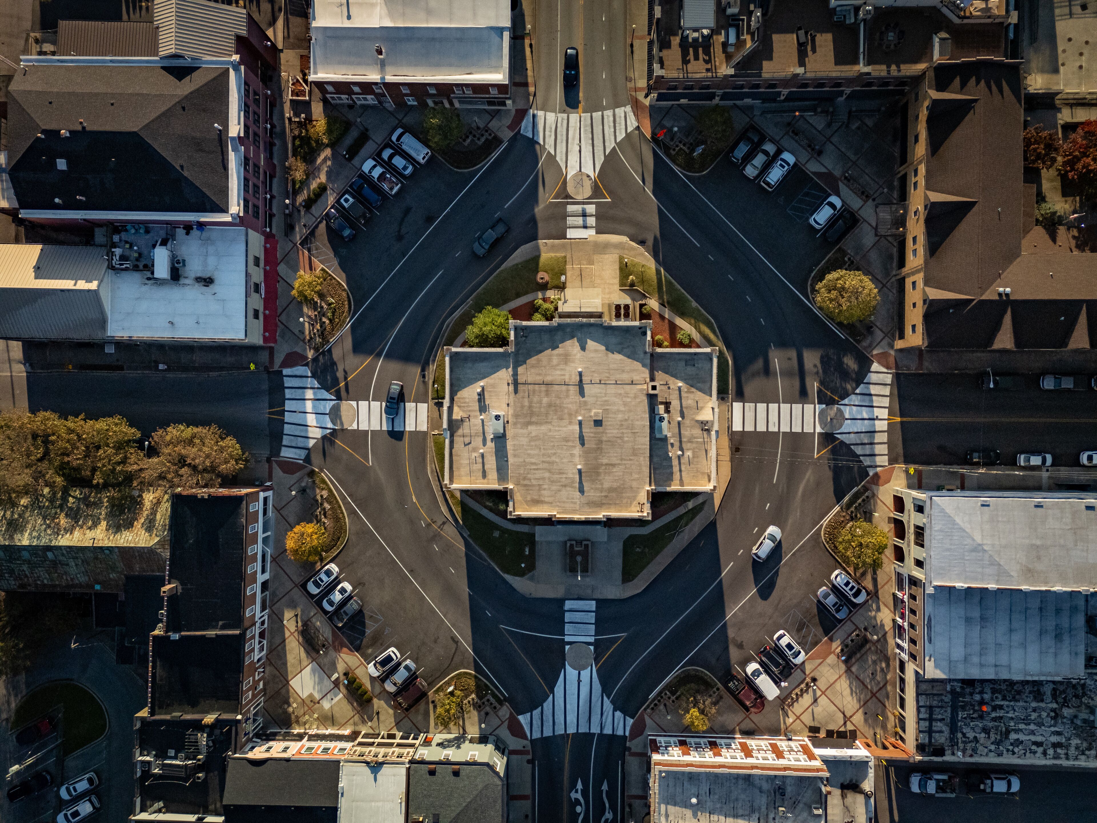An aerial view of a roundabout with a central plaza, surrounded by buildings and parked cars.