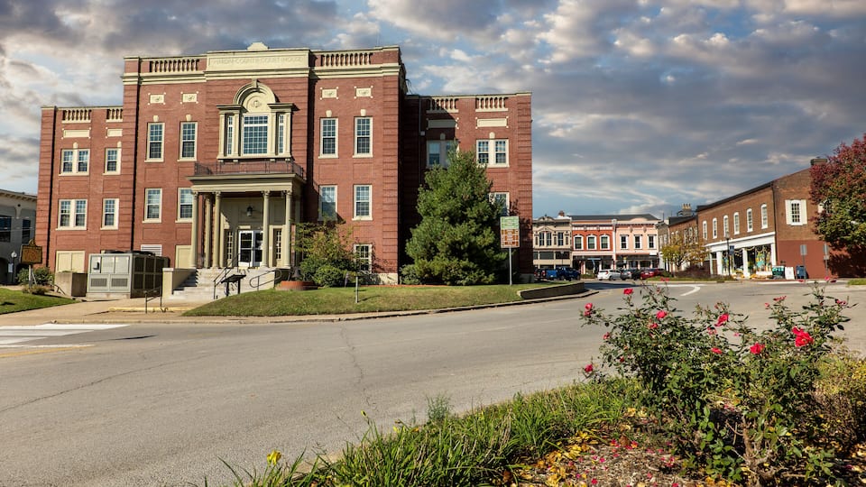 A historic courthouse with a red brick facade, surrounded by a street with parked cars and flowering bushes.